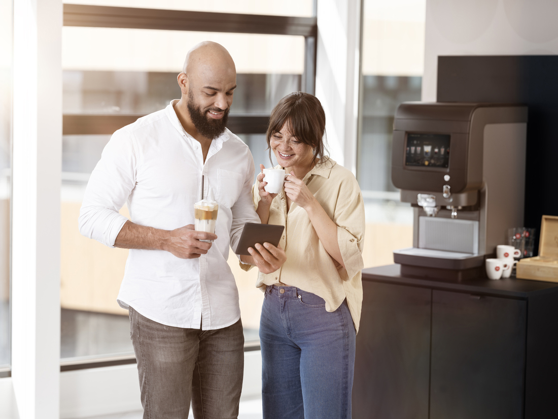 Deux personnes échangent près d’une machine à café moderne, tablette en main et boissons à la main.