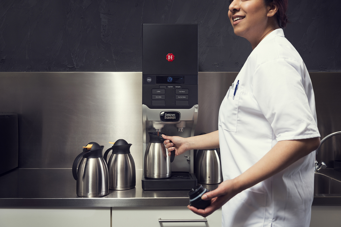 Un professionnel de santé en uniforme blanc remplit une cafetière à une machine à café Douwe Egberts dans une salle de pause.