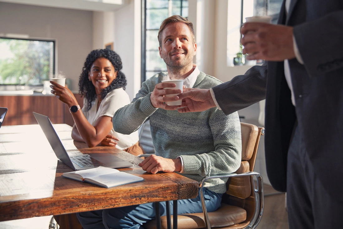 Trois collègues échangent autour d’une table de réunion, café à la main dans un cadre professionnel.